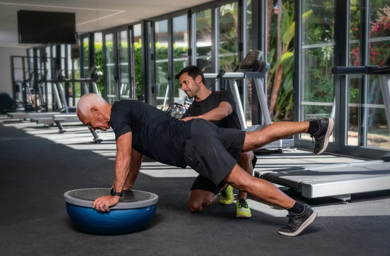 An older adult practicing a single-leg balance drill with a coach nearby