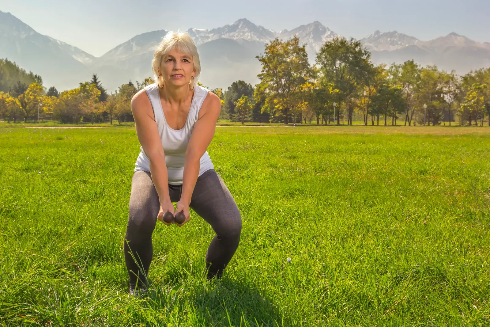 Older woman doing squats with dumbbells in a park with mountains behind, a strength lift for women over 50