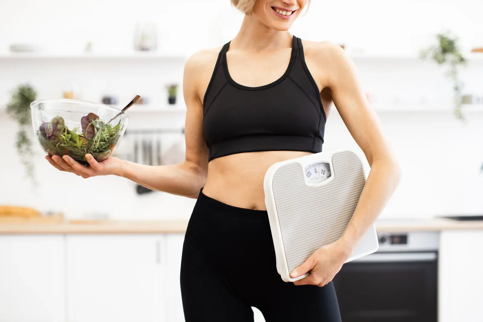 Fit woman holding a salad bowl and a bathroom scale in the kitchen, balancing a calorie deficit with training