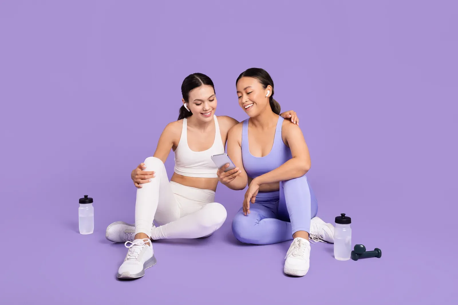Two women checking a phone during a fitness break with water bottles, staying hydrated while on a GLP-1