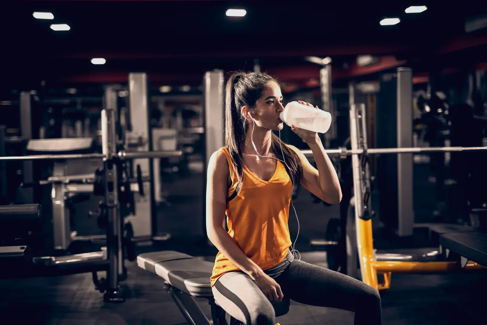Woman with headphones drinking water while sitting on a bench in the gym, a pause that can help with dizziness