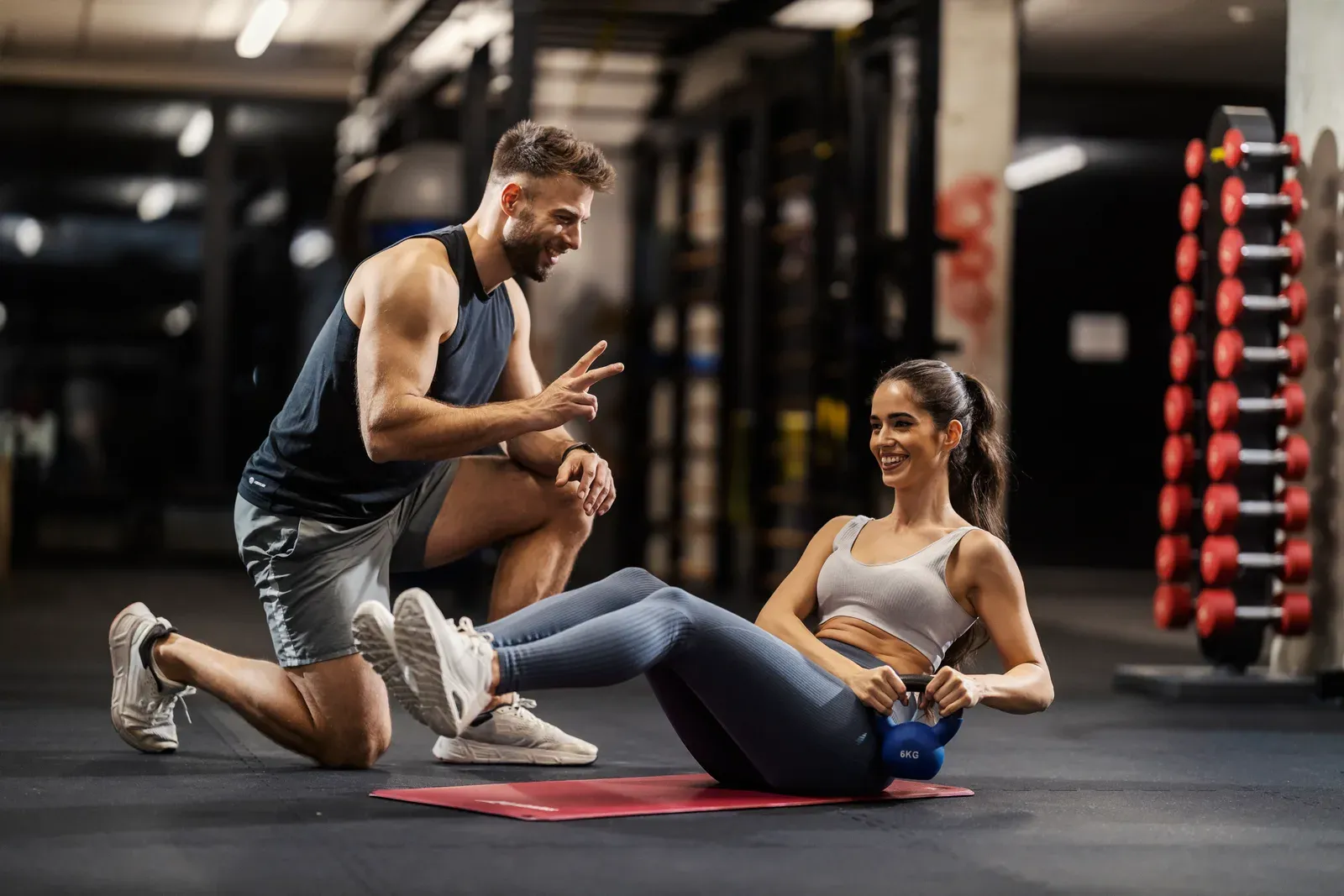 Sportswoman doing a Russian twist with a kettlebell in a gym while her trainer kneels beside her, a trainer in action