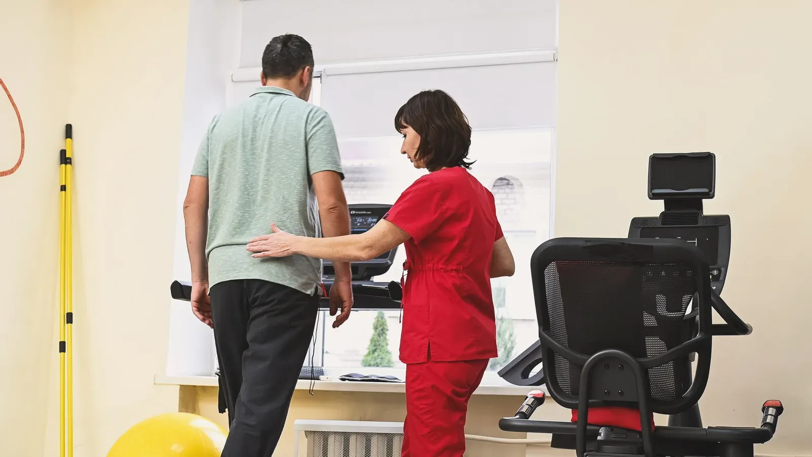 Middle-aged man exercising on a treadmill with a physical therapist while catching a ball, guided clinical-style training