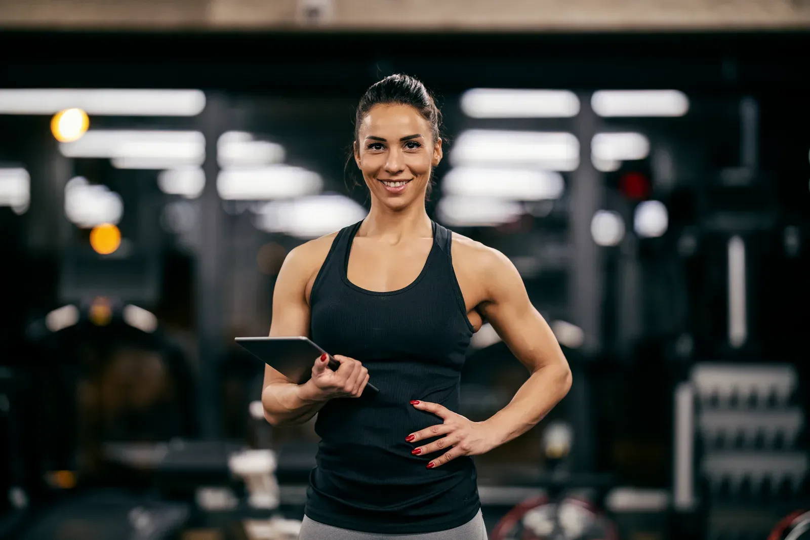Fit muscular female personal trainer holding a tablet and smiling at the camera in a gym