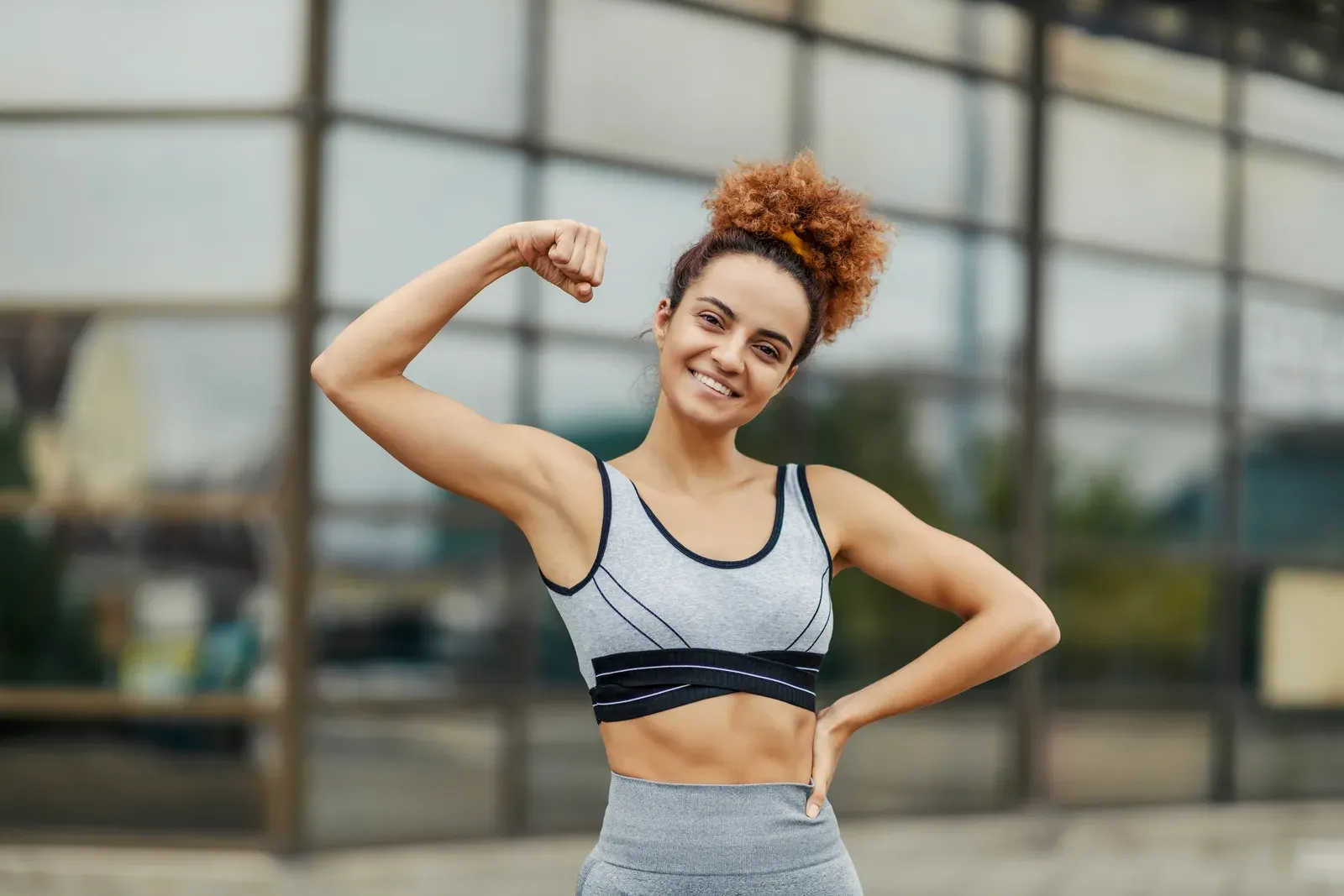 Happy strong sportswoman showing her biceps while standing outdoors, a picture of muscle definition for women