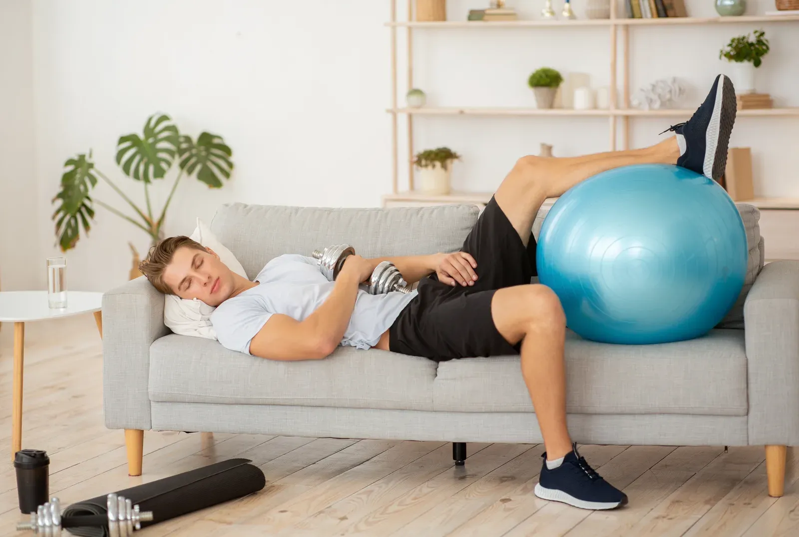 Tired man asleep on a couch with a dumbbell and fitness ball, the downside of stopping training