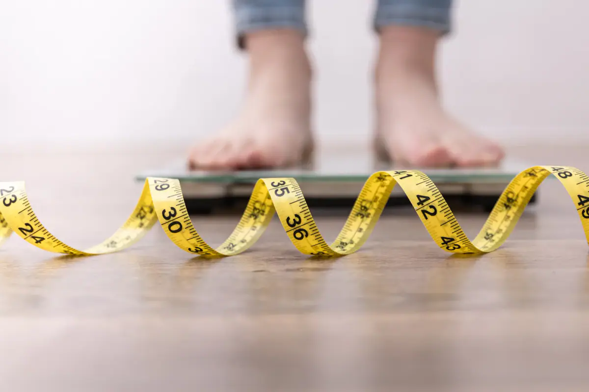 Stepping onto a bathroom scale beside a tape measure to check weight loss