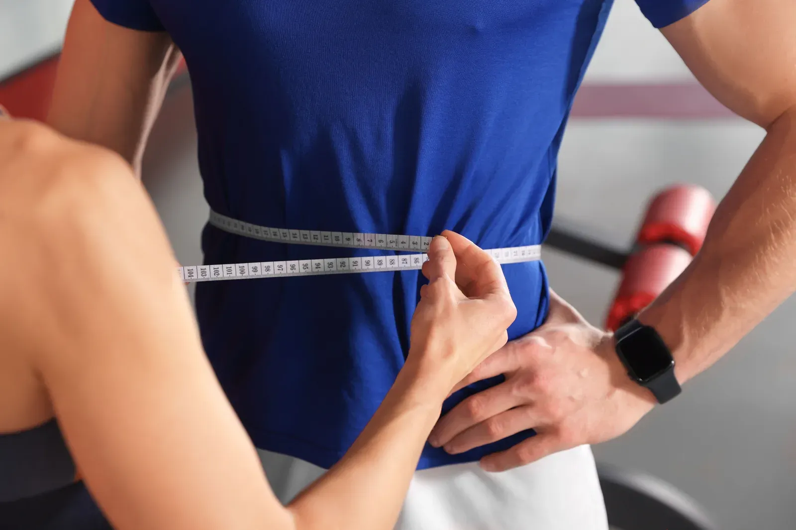 Trainer measuring a man's waist with a tape in the gym, a way to track a beer belly