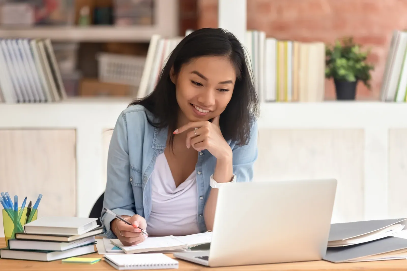 Smiling student studying in a library with a laptop and books, as when researching an online trainer certification